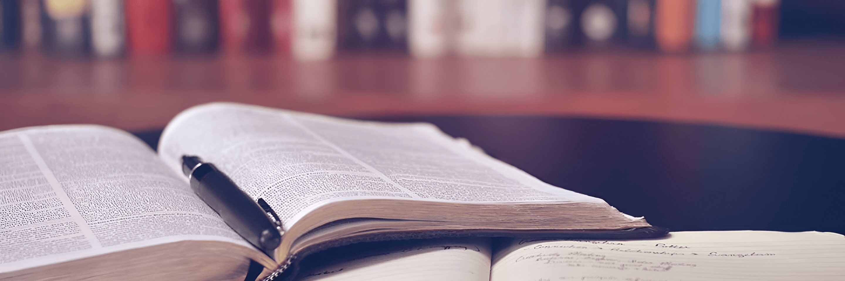 An open book with a pen on top is placed on a desk next to a partially visible notebook. Shelves filled with books are blurred in the background.