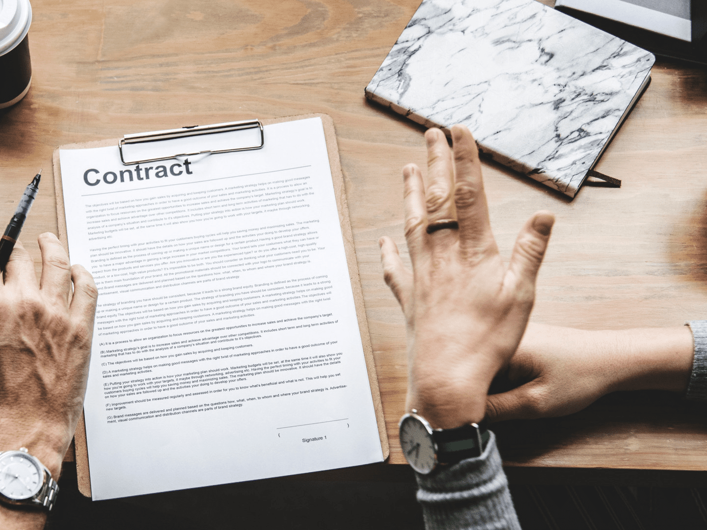 Two people discussing a contract on a clipboard at a wooden table, with a notebook and pen nearby.